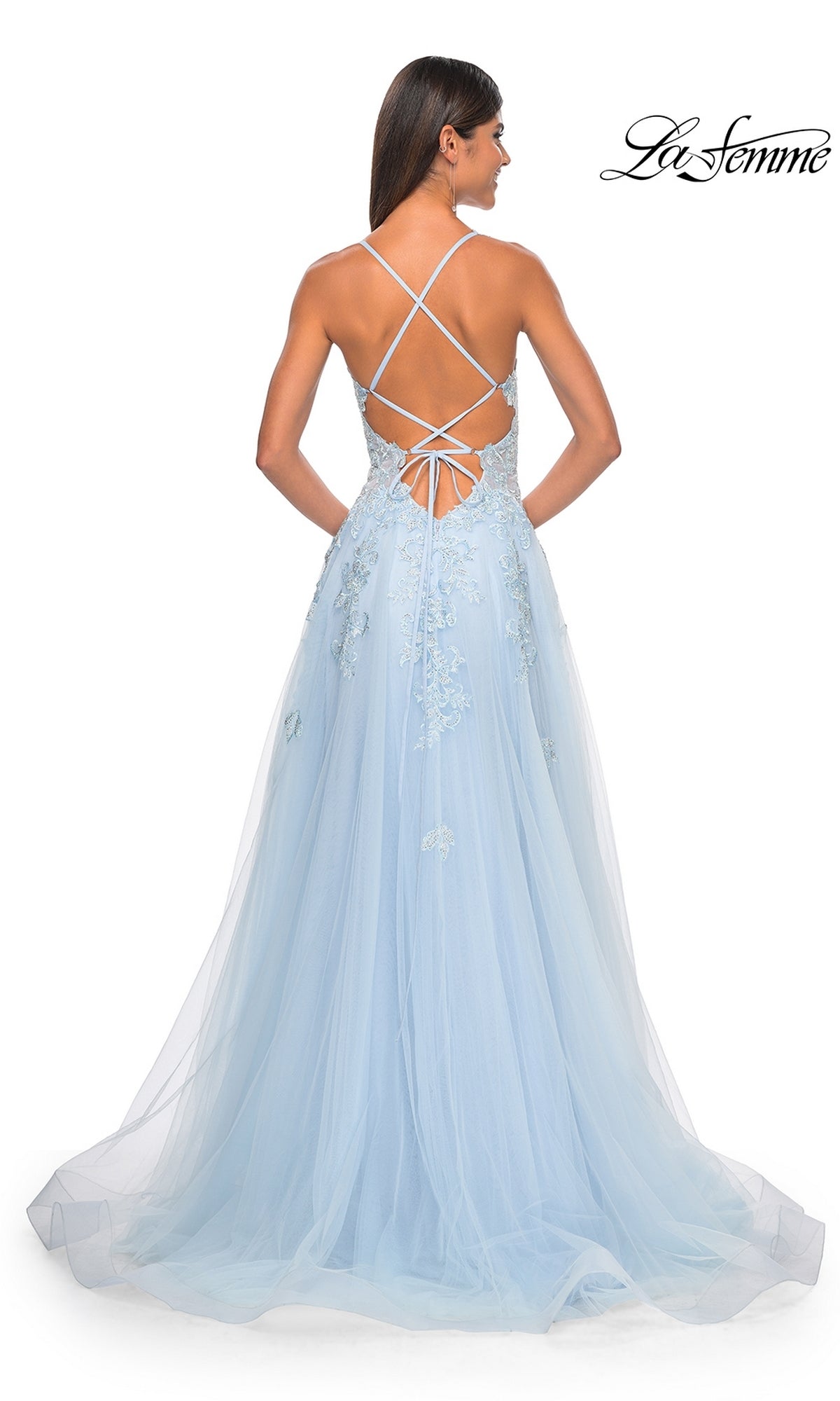 A woman in the Sheer-Waist Long A-Line Prom Dress (La Femme 32438) stands facing away. The light blue dress features delicate lace embroidery, a strappy open back, and a flowing tulle skirt.